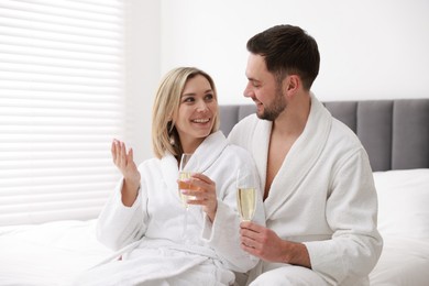 Photo of Spa. Happy couple in bathrobes with glasses of champagne sitting on bed indoors