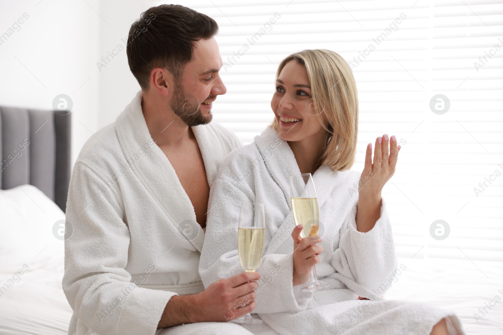 Photo of Spa. Happy couple in bathrobes with glasses of champagne sitting on bed indoors