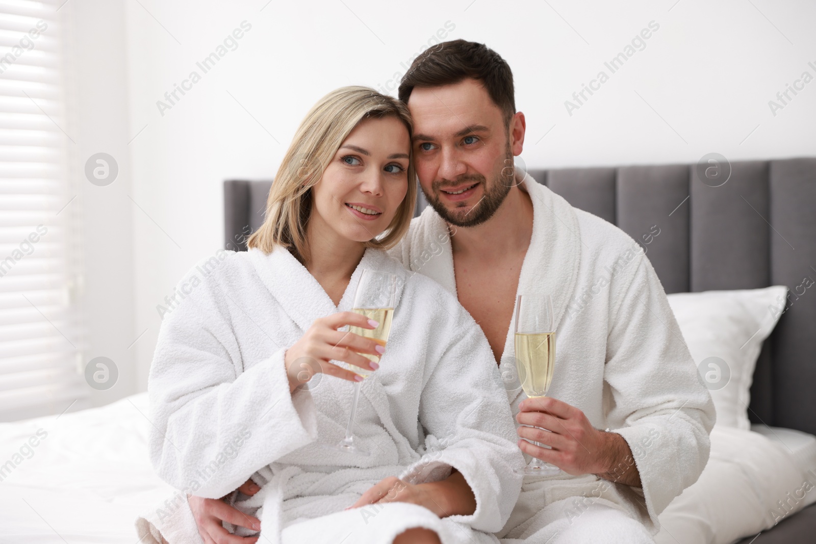 Photo of Spa. Happy couple in bathrobes with glasses of champagne sitting on bed indoors