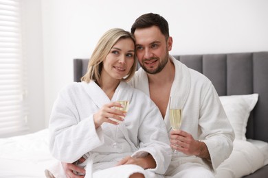 Photo of Spa. Happy couple in bathrobes with glasses of champagne sitting on bed indoors