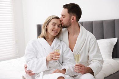 Photo of Spa. Happy couple in bathrobes with glasses of champagne sitting on bed indoors