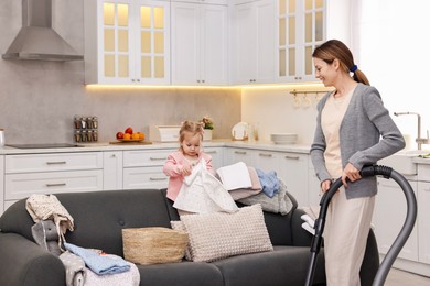 Photo of Smiling housewife cleaning with vacuum cleaner near sofa and her little daughter at home