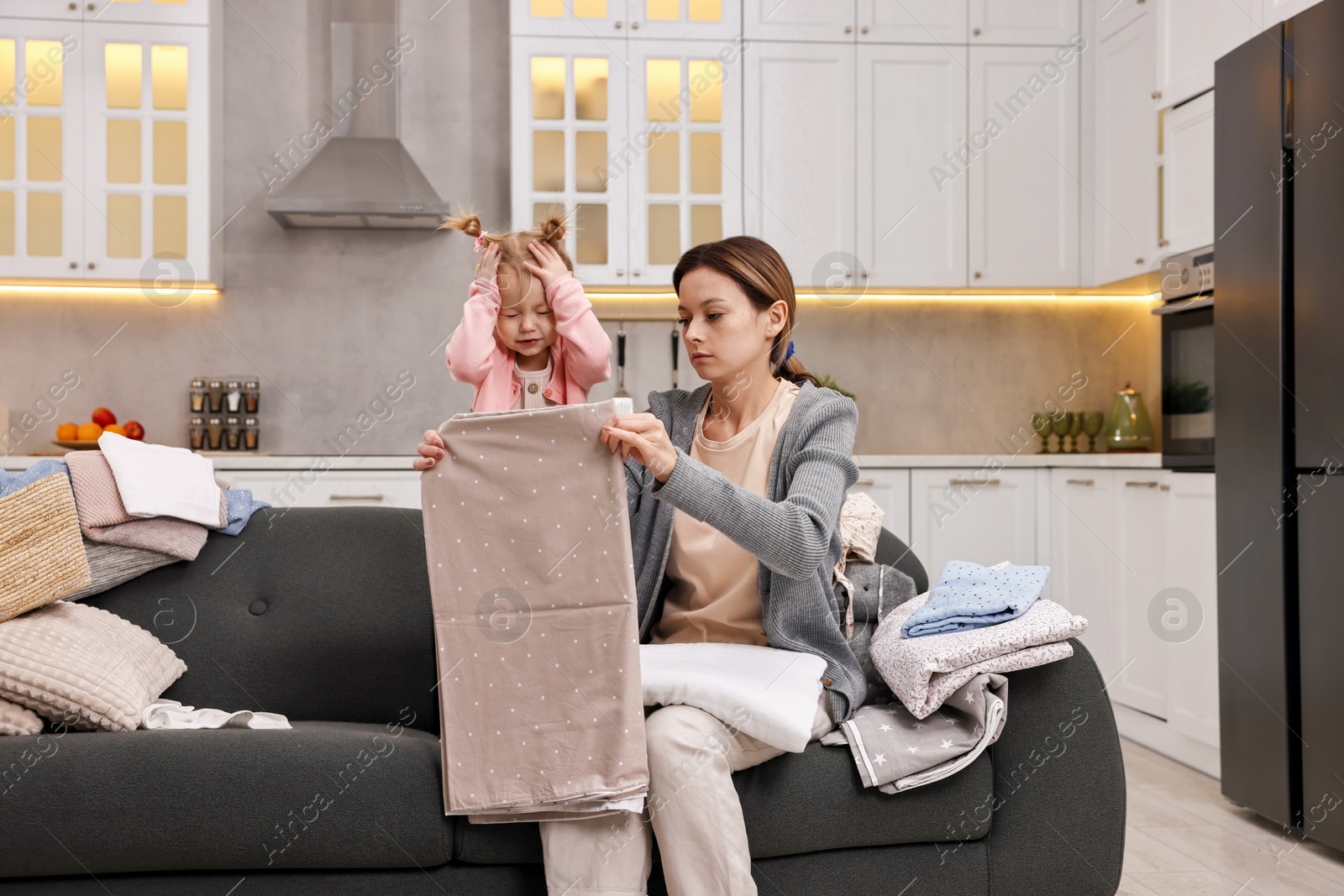 Photo of Housewife with her little daughter folding laundry on sofa at home