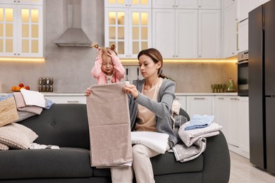 Photo of Housewife with her little daughter folding laundry on sofa at home