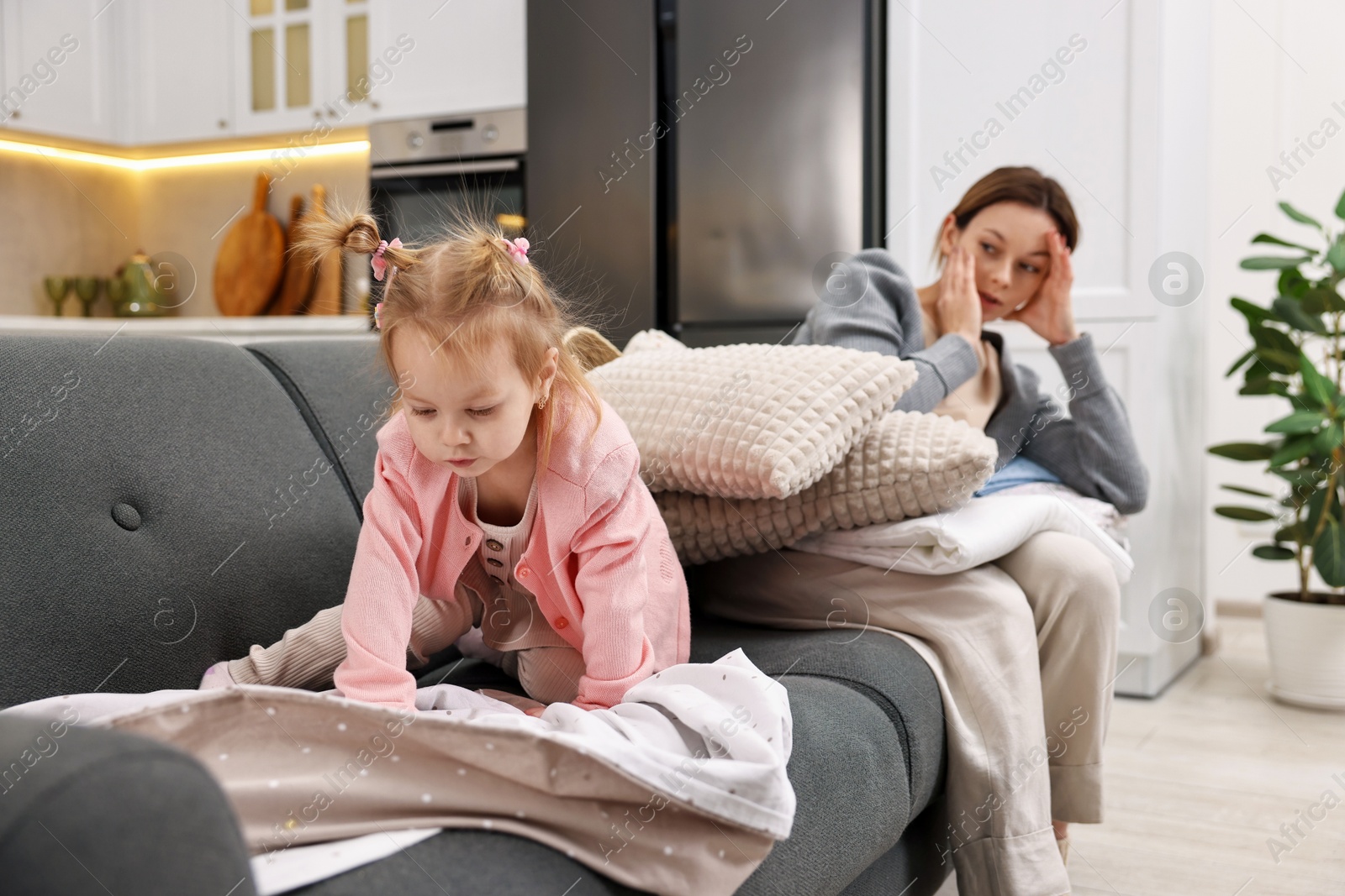 Photo of Tired housewife with her little daughter among messy laundry on sofa at home