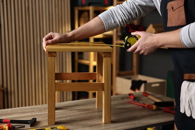 Photo of Repairman measuring wooden stool at table in workshop, closeup