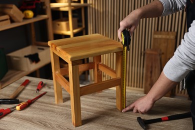 Photo of Repairman measuring wooden stool at table in workshop, closeup