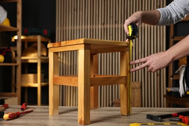 Photo of Repairman measuring wooden stool at table in workshop, closeup