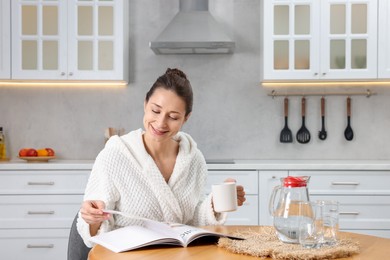 Photo of Beautiful woman with cup of drink reading magazine after spa procedure at wooden table in kitchen