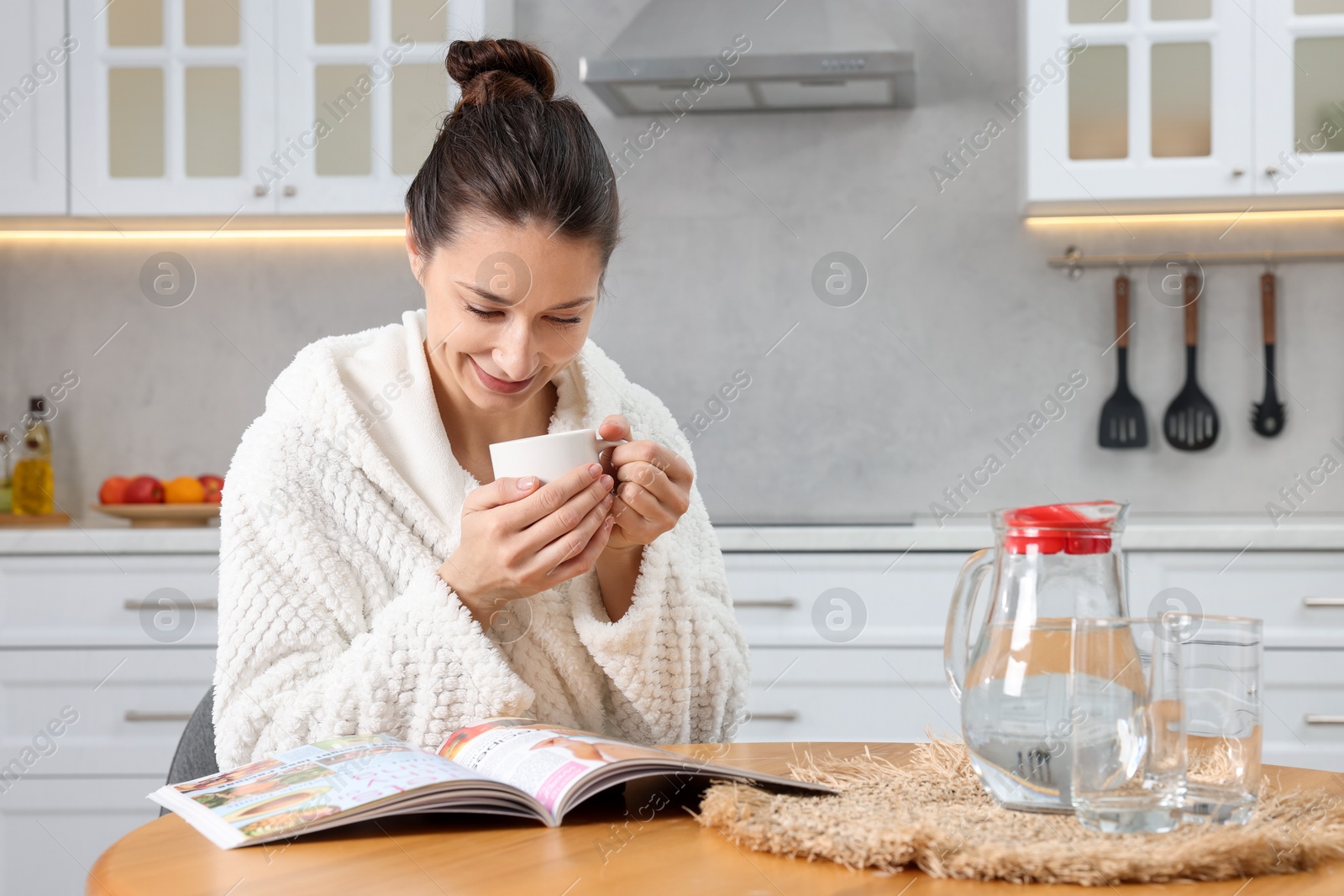 Photo of Beautiful woman with cup of drink reading magazine after spa procedure at wooden table in kitchen