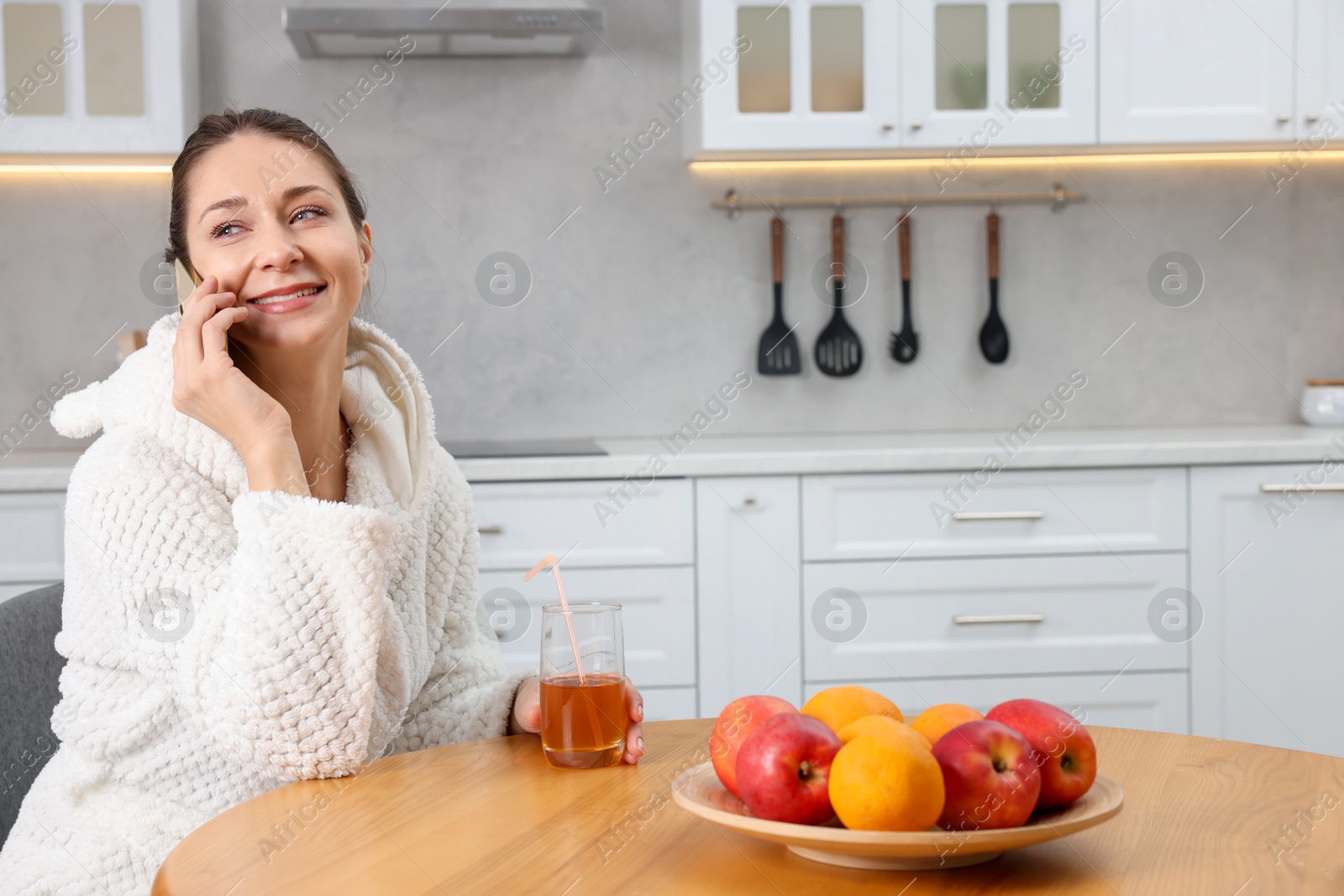 Photo of Beautiful woman with glass of juice talking on smartphone after spa procedure in kitchen