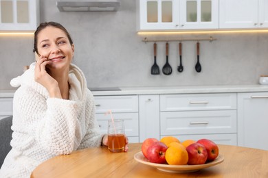 Photo of Beautiful woman with glass of juice talking on smartphone after spa procedure in kitchen