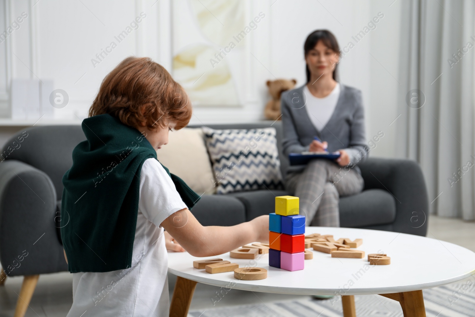 Photo of Boy playing with wooden letters at table while psychologist taking notes, selective focus