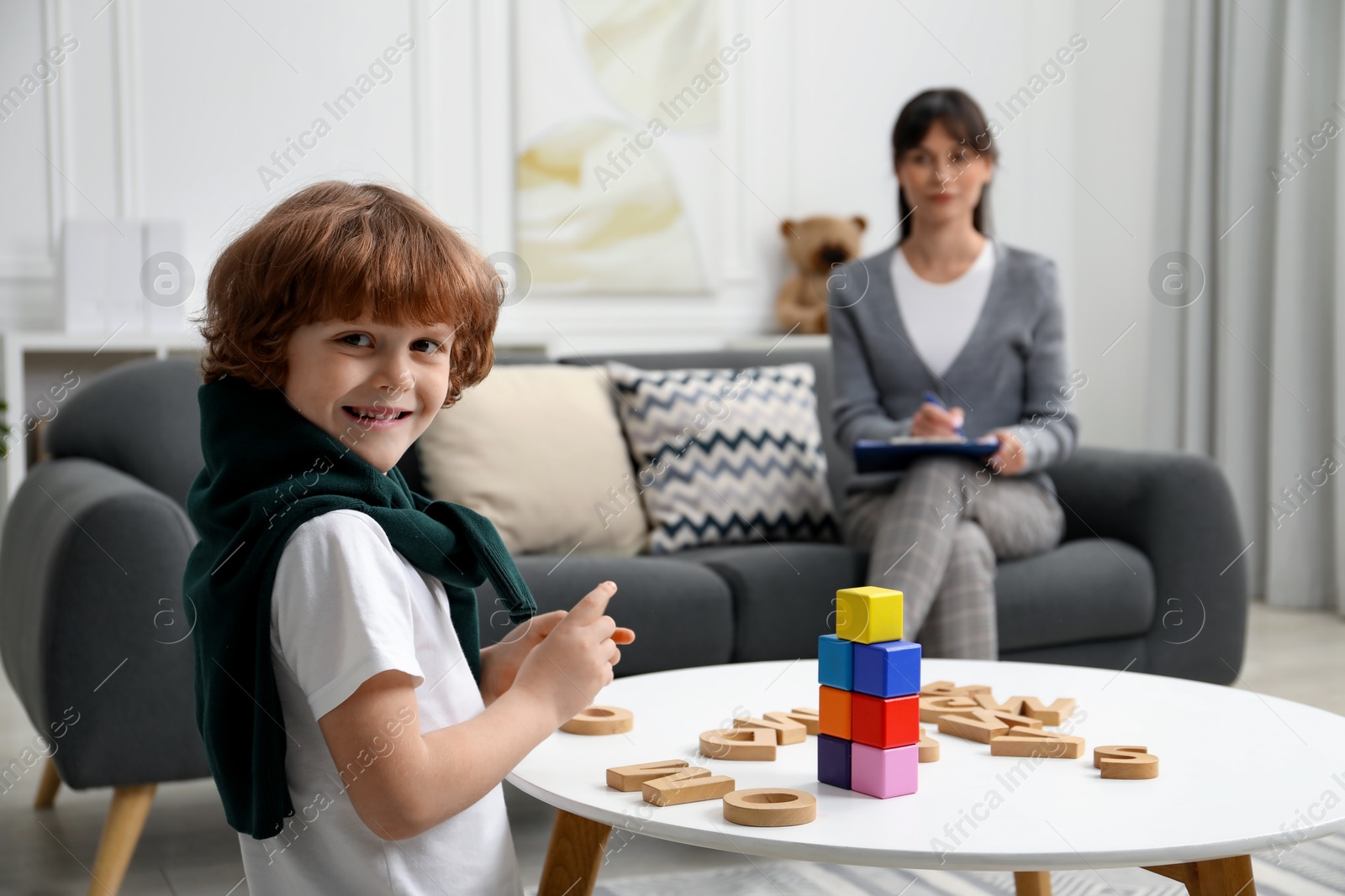 Photo of Boy playing with wooden letters at table while psychologist taking notes, selective focus