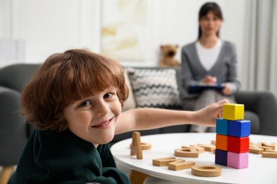 Photo of Boy playing with wooden cubes at table while psychologist taking notes, selective focus