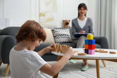 Photo of Boy playing with wooden letters at table while psychologist taking notes, selective focus