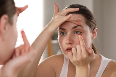 Photo of Young woman putting in red color contact lens near mirror indoors