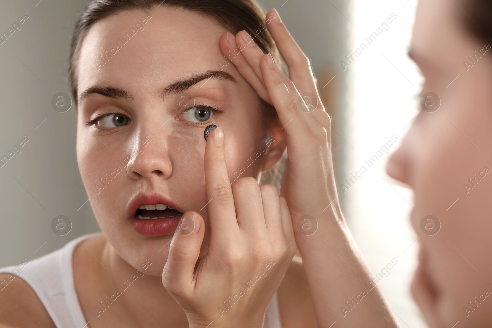 Photo of Young woman putting in blue color contact lens near mirror indoors, closeup