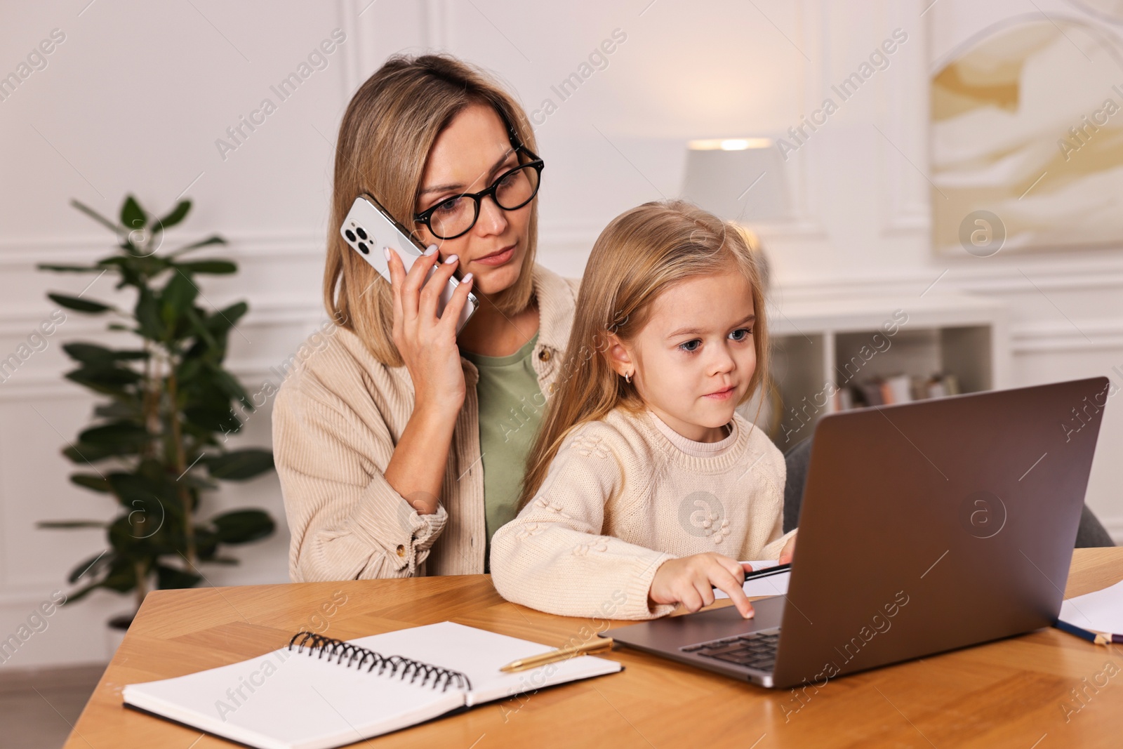 Photo of Work-family balance. Single mother talking by smartphone and her daughter drawing at wooden table indoors