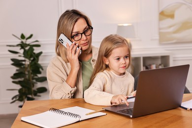 Work-family balance. Single mother talking by smartphone and her daughter drawing at wooden table indoors