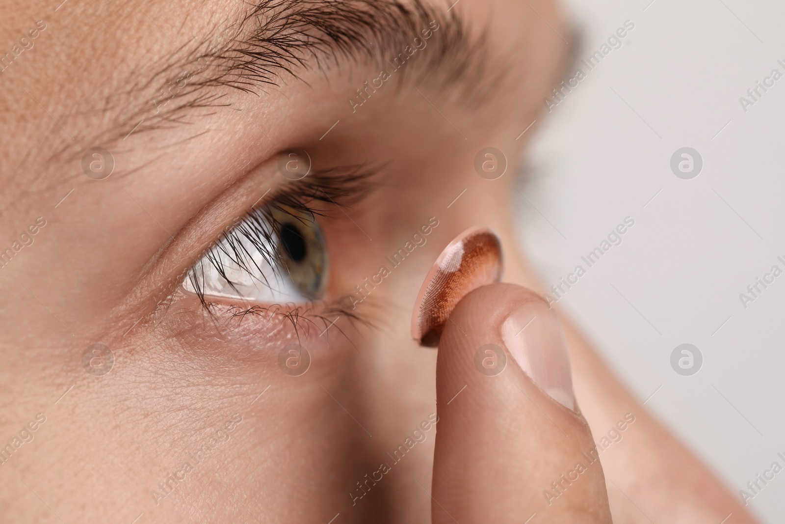 Photo of Man putting color contact lens in his eye on light background, closeup