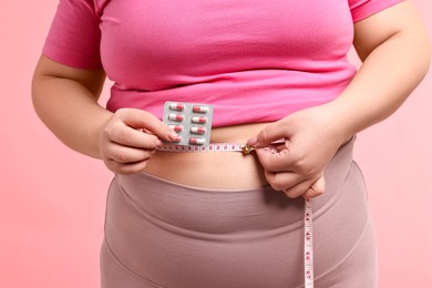 Photo of Plus size woman measuring waist with tape and holding blister of weight loss supplements on pink background, closeup