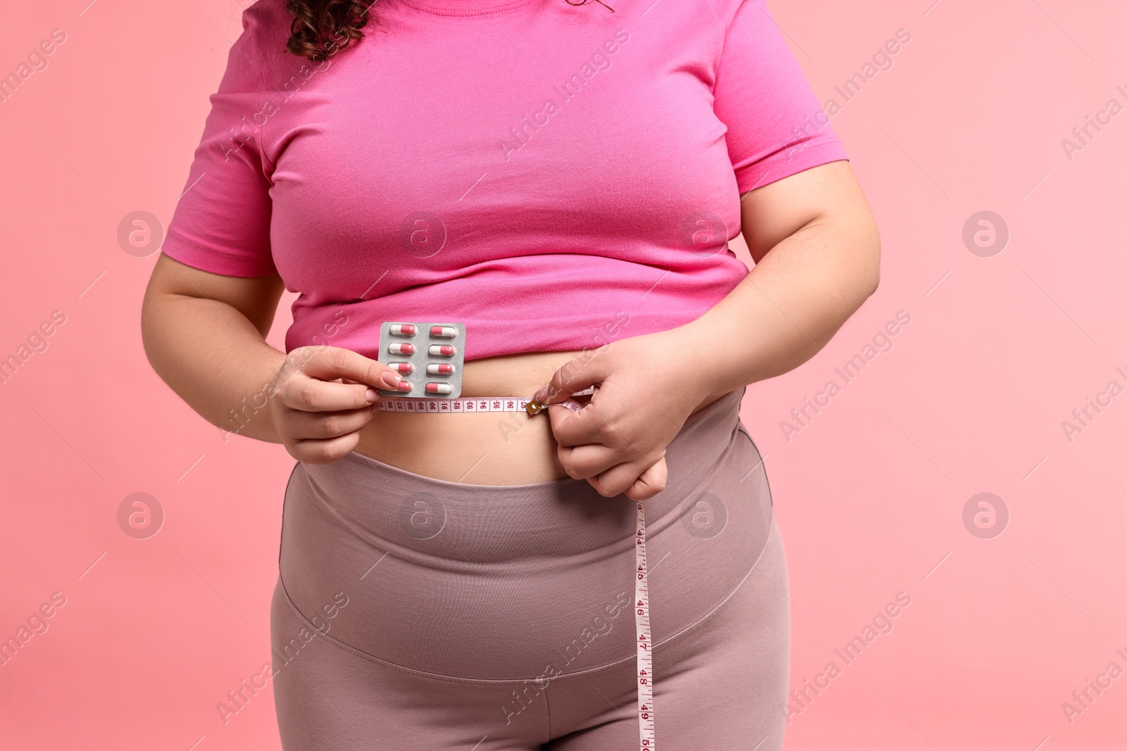 Photo of Plus size woman measuring waist with tape and holding blister of weight loss supplements on pink background, closeup