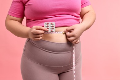 Photo of Plus size woman measuring waist with tape and holding blister of weight loss supplements on pink background, closeup