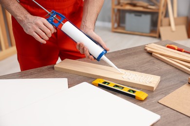 Photo of Worker with caulking gun glueing wooden plank indoors, closeup