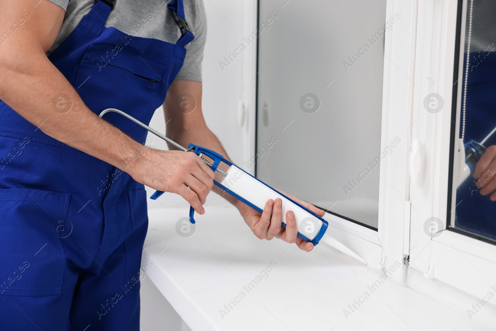 Photo of Worker with caulking gun sealing window indoors, closeup