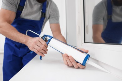 Photo of Worker with caulking gun sealing window indoors, closeup