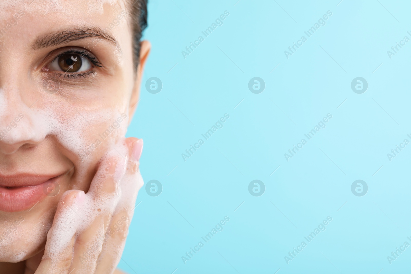 Photo of Attractive woman washing her face with cleansing foam on light blue background, closeup. Space for text