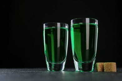 Photo of Absinthe in shot glasses and brown sugar on table against black background, space for text