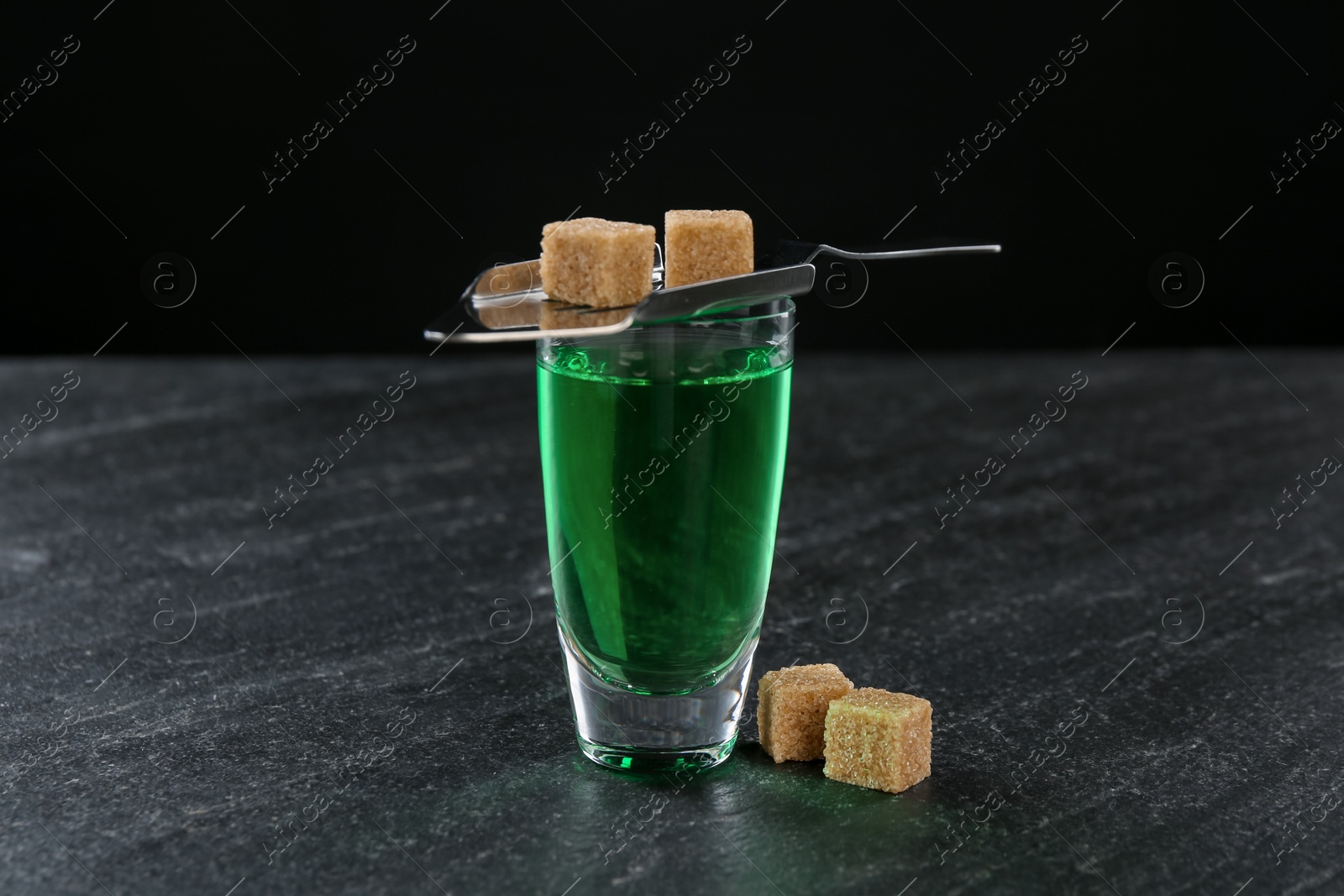 Photo of Absinthe in shot glass, brown sugar and slotted spoon on black table, closeup