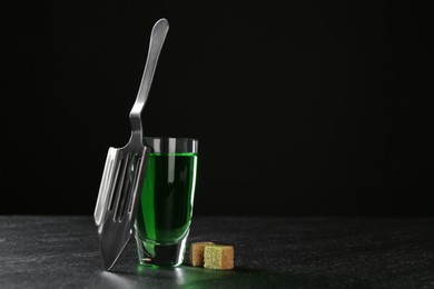 Photo of Absinthe in shot glass, brown sugar and slotted spoon on table against black background, space for text