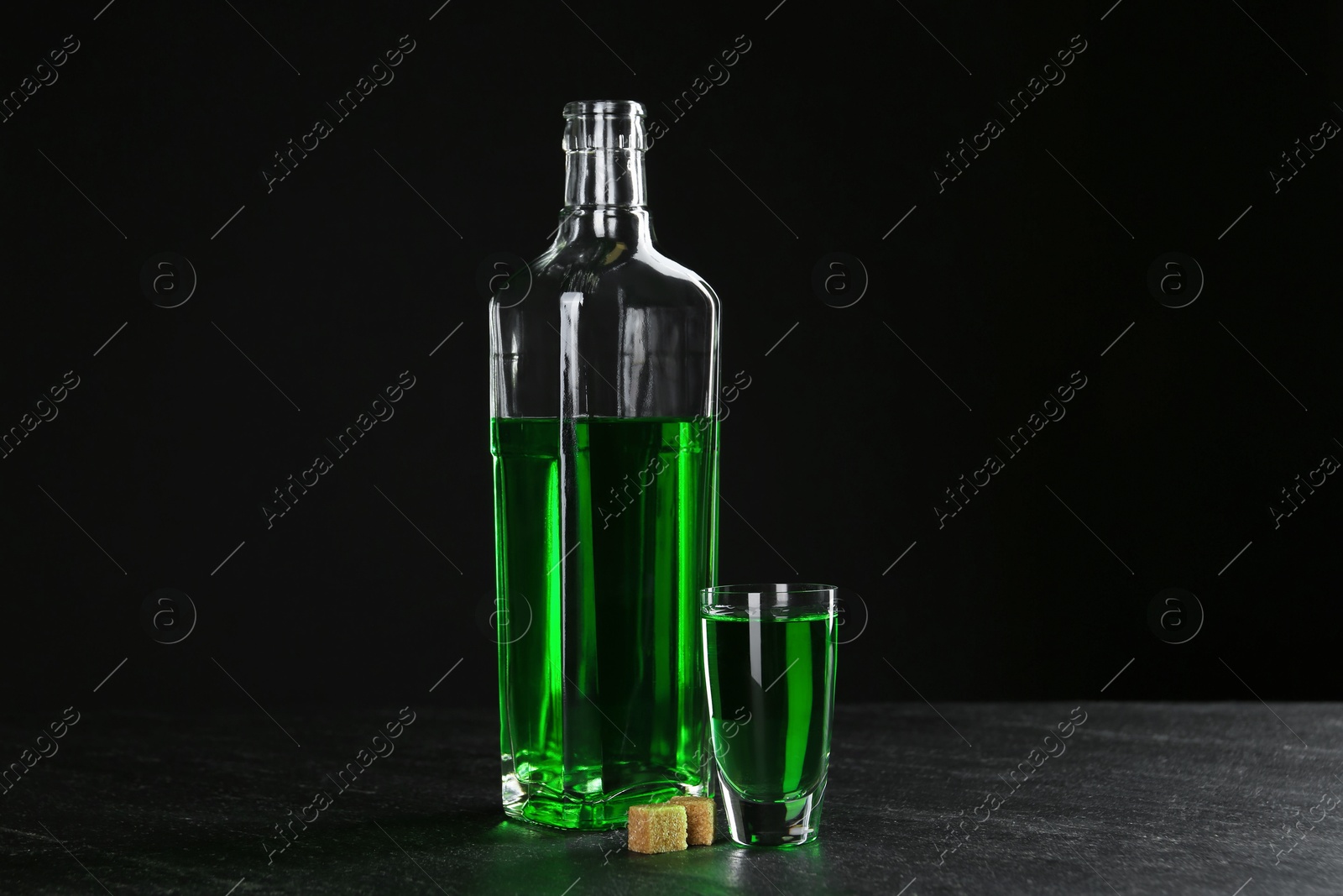 Photo of Absinthe in shot glass, bottle and brown sugar on table against black background