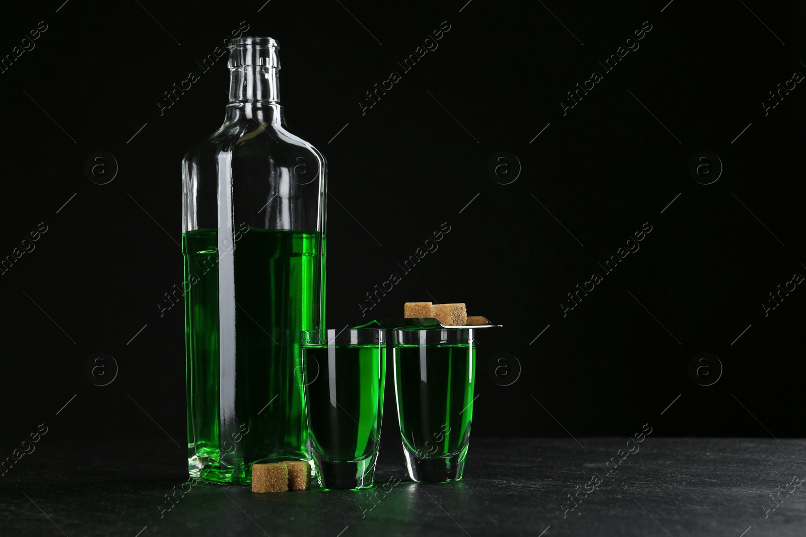 Photo of Absinthe in shot glasses, bottle, brown sugar and slotted spoon on table against black background, space for text