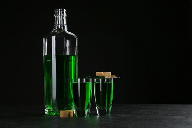 Photo of Absinthe in shot glasses, bottle, brown sugar and slotted spoon on table against black background, space for text