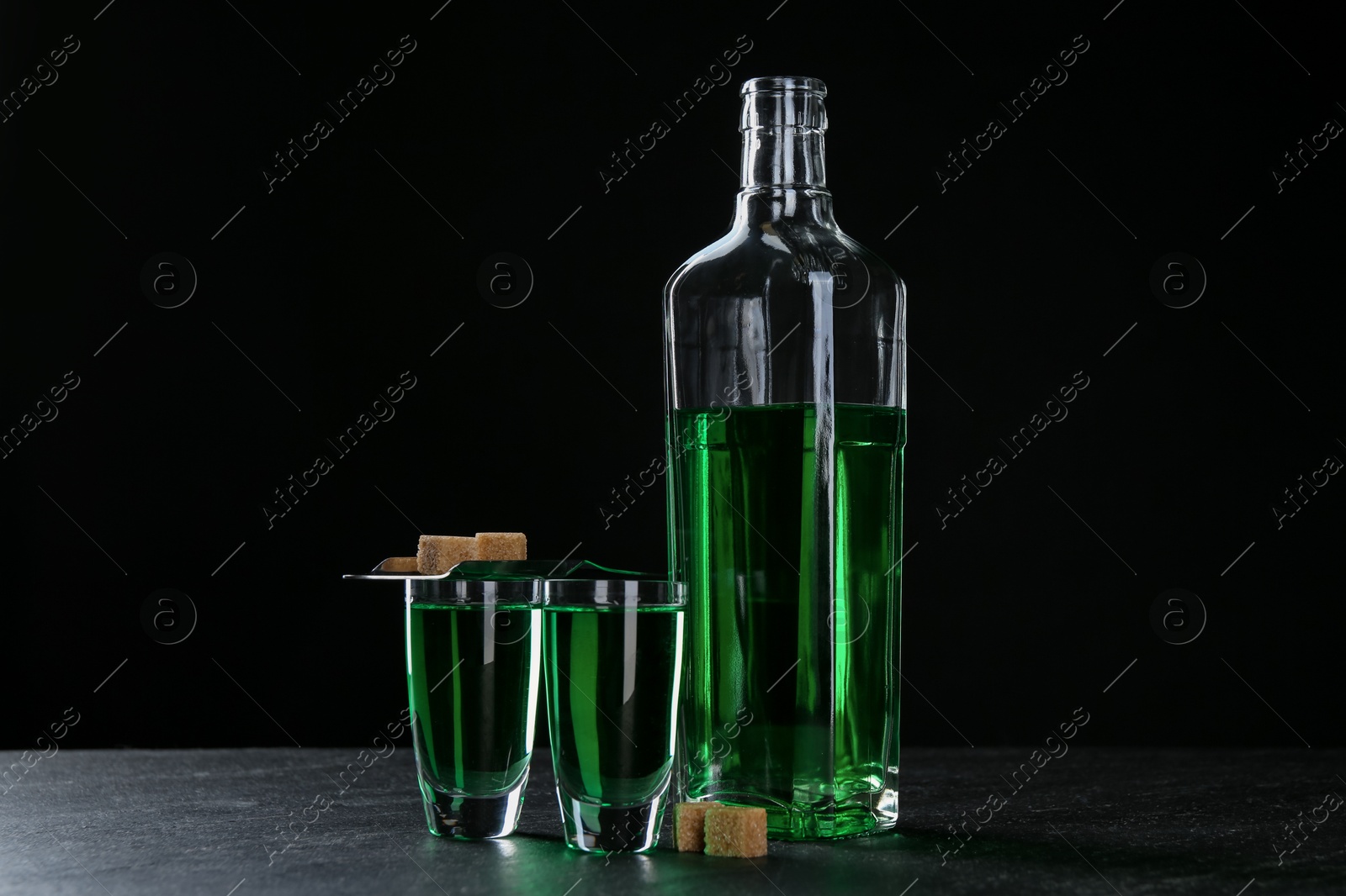 Photo of Absinthe in shot glasses, bottle, brown sugar and slotted spoon on table against black background