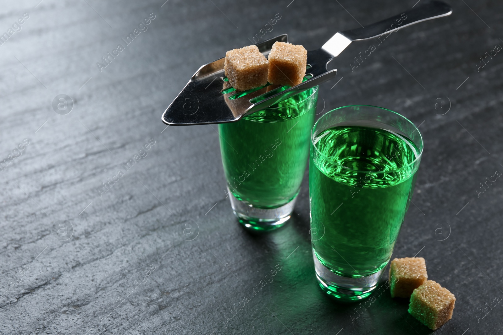 Photo of Absinthe in shot glasses, brown sugar and slotted spoon on black table, closeup. Space for text