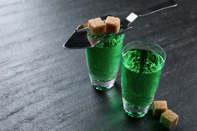 Photo of Absinthe in shot glasses, brown sugar and slotted spoon on black table, closeup. Space for text