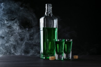 Photo of Absinthe in shot glasses, bottle and brown sugar on table against black background with smoke