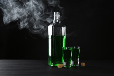 Photo of Absinthe in shot glasses, bottle and brown sugar on table against black background with smoke