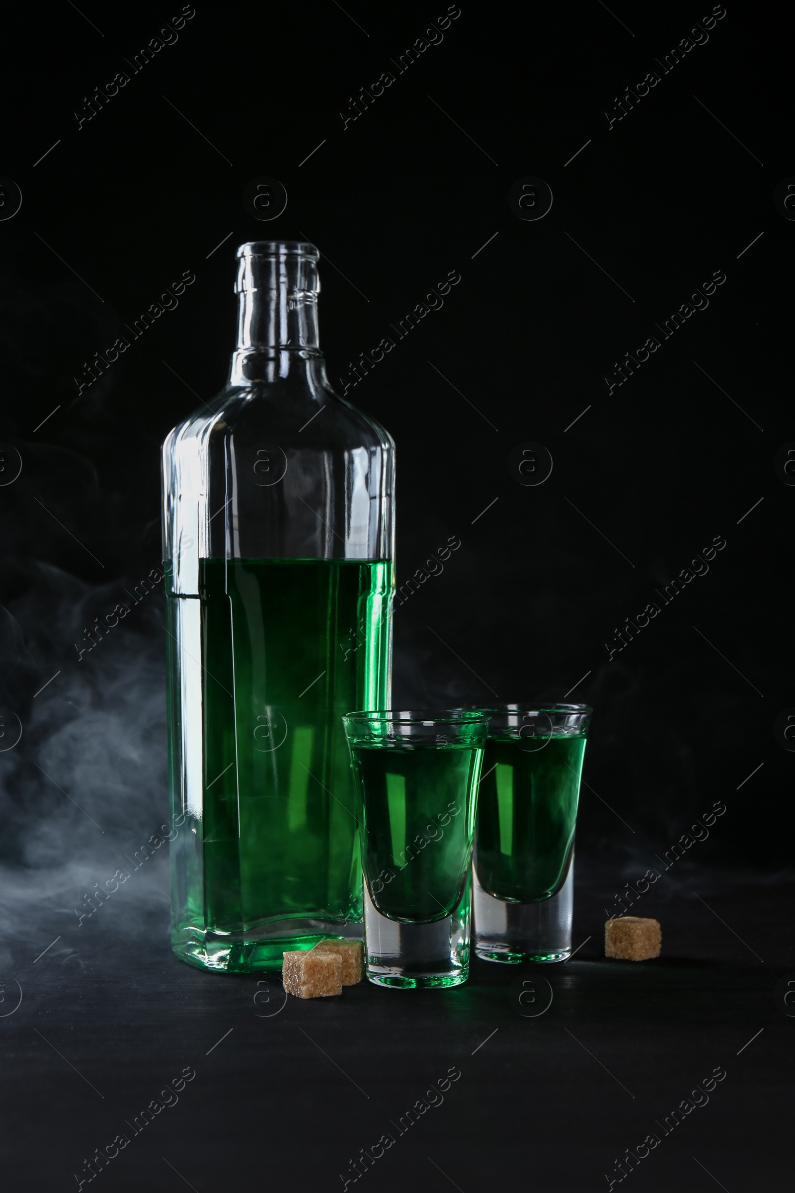 Photo of Absinthe in shot glasses, bottle and brown sugar on table against black background with smoke