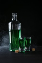 Photo of Absinthe in shot glasses, bottle and brown sugar on table against black background with smoke