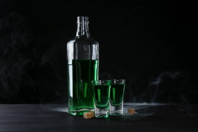 Photo of Absinthe in shot glasses, bottle and brown sugar on table against black background with smoke