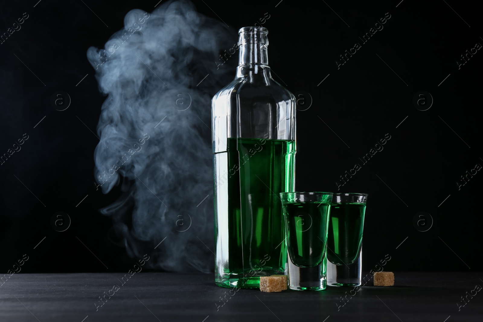 Photo of Absinthe in shot glasses, bottle and brown sugar on table against black background with smoke, space for text