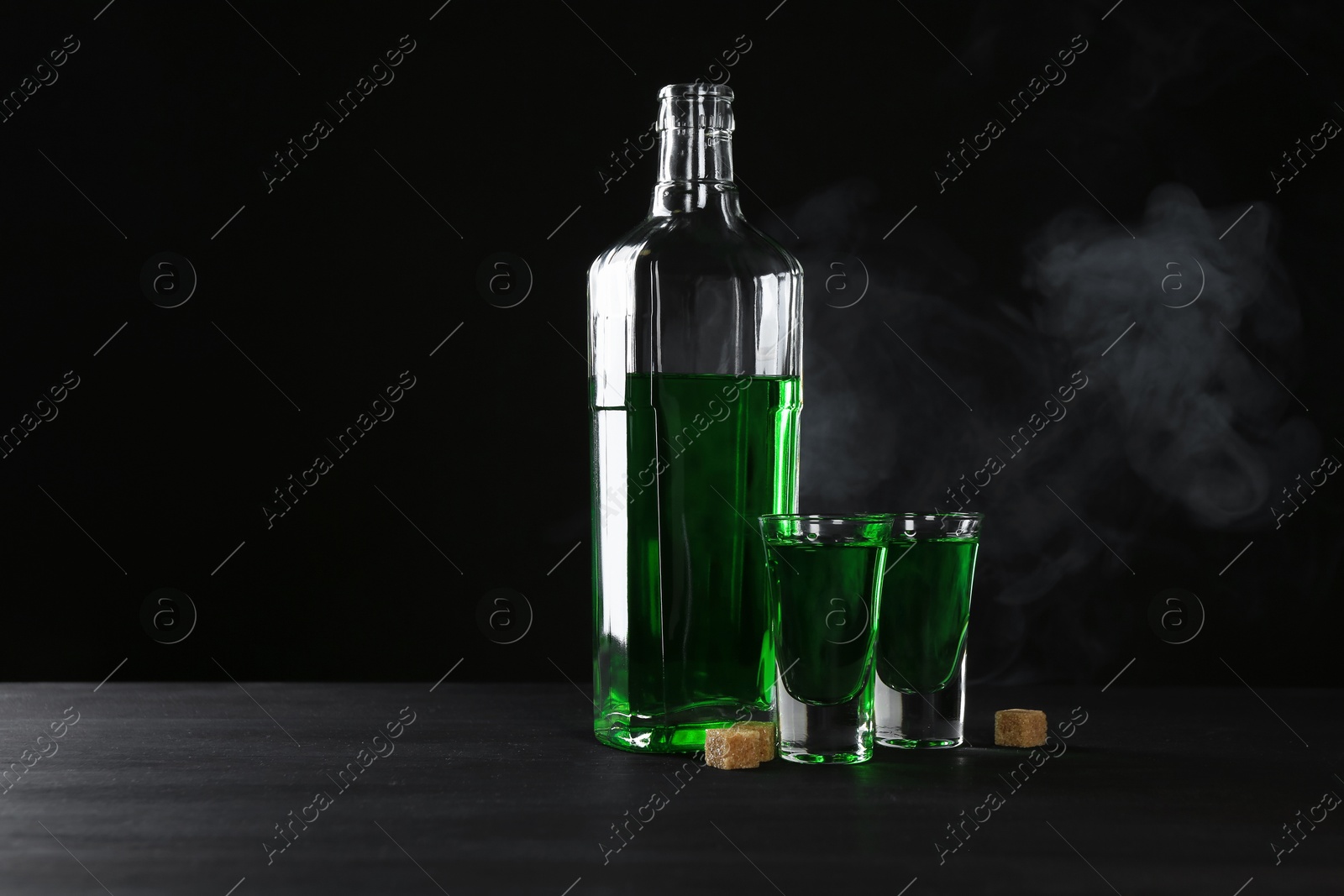 Photo of Absinthe in shot glasses, bottle and brown sugar on table against black background with smoke, space for text