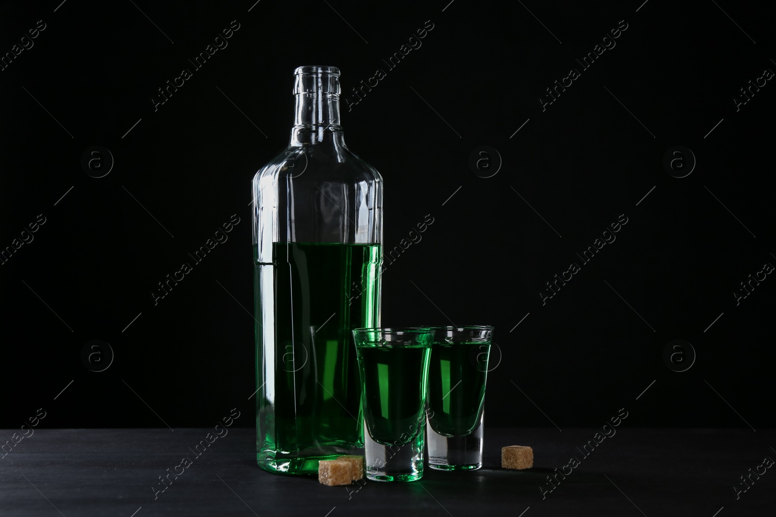 Photo of Absinthe in shot glasses, bottle and brown sugar on table against black background