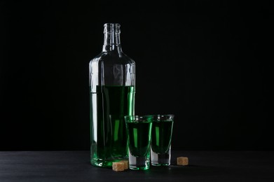 Photo of Absinthe in shot glasses, bottle and brown sugar on table against black background